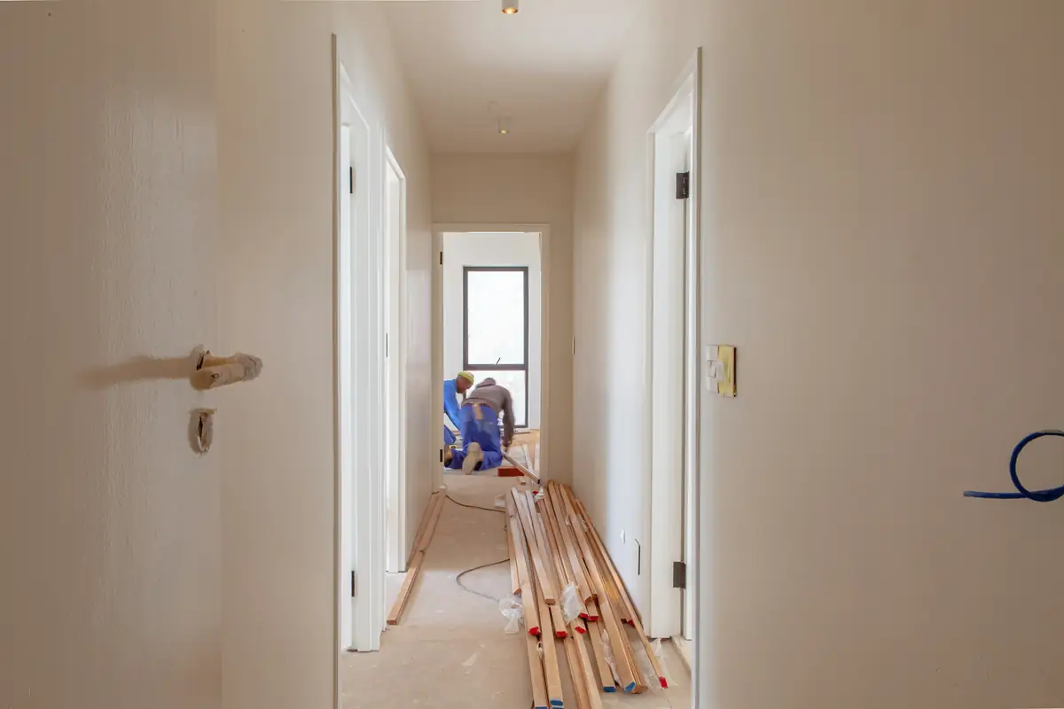 Couloir en travaux : murs repeints, tasseaux au sol et deux artisans au fond