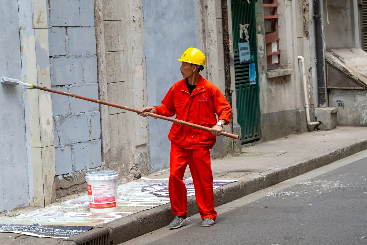 Peintre en tenue de travail passant un rouleau à perche sur un mur, bâche et pot au sol